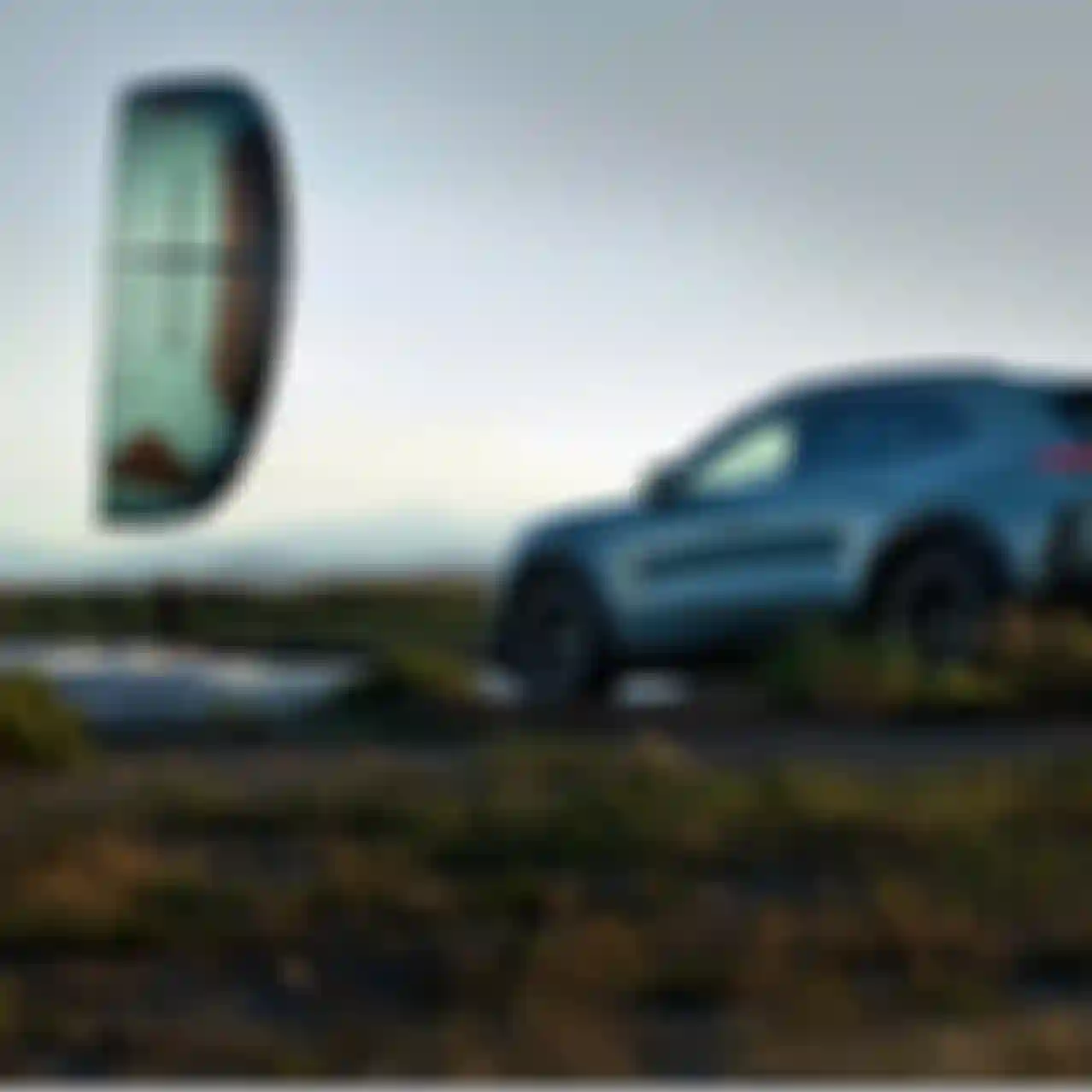 A kite surfer rides the waves near a sleek, parked SUV on a grassy shore during sunset, with mountains visible in the background.