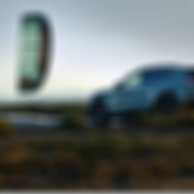 A kite surfer rides the waves near a sleek, parked SUV on a grassy shore during sunset, with mountains visible in the background.