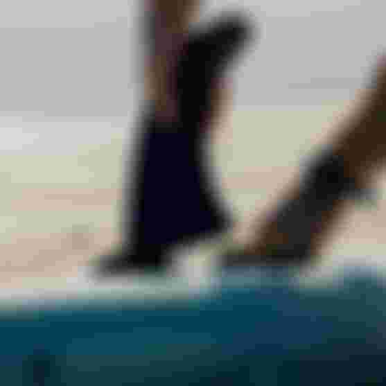 Hands attaching a fin to a surfboard on a sandy beach, with a person wearing a wristwatch.