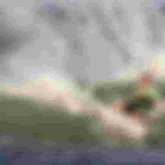 James Carew kite surfing on a wave, creating a large splash under a clear blue sky.