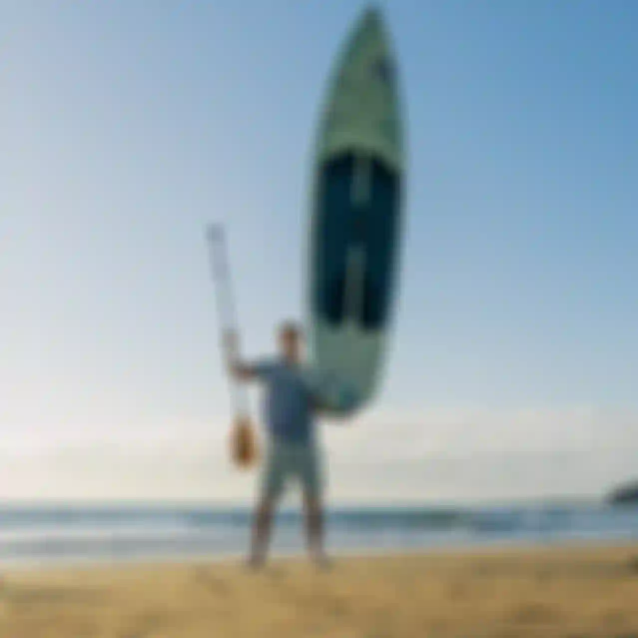 Person on a sandy beach holding a paddleboard vertically and a paddle, with the sea and a distant island in the background under a clear sky.