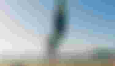 Person on a sandy beach holding a paddleboard vertically and a paddle, with the sea and a distant island in the background under a clear sky.