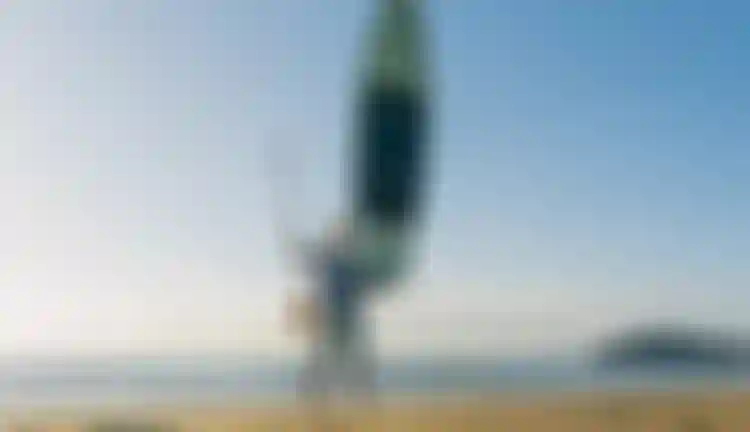 Person on a sandy beach holding a paddleboard vertically and a paddle, with the sea and a distant island in the background under a clear sky.