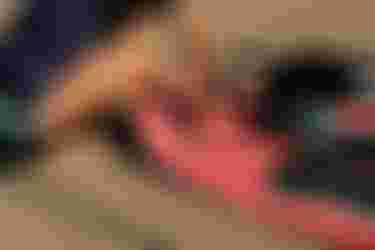 Person kneeling on sand inflating a red paddleboard with a pump. Wearing a dark shirt and turquoise shorts, hands adjusting the nozzle.