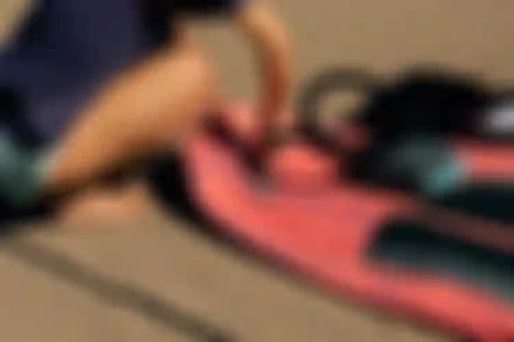 Person kneeling on sand inflating a red paddleboard with a pump. Wearing a dark shirt and turquoise shorts, hands adjusting the nozzle.