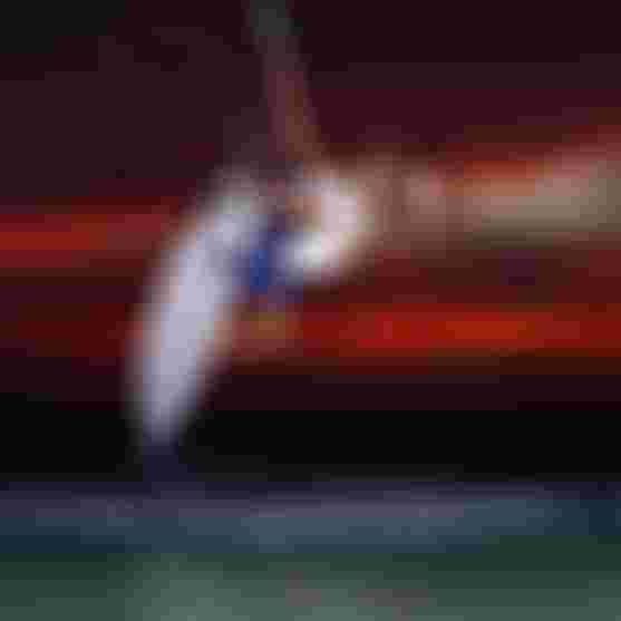 Male kitesurfer mid-air above ocean waves at dramatic red sunset, gripping kite lines while holding white surfboard vertically