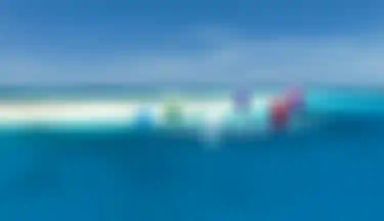 Aerial view of multiple colorful kites and kitesurfers riding turquoise tropical waters near a white sand reef under clear blue sky.