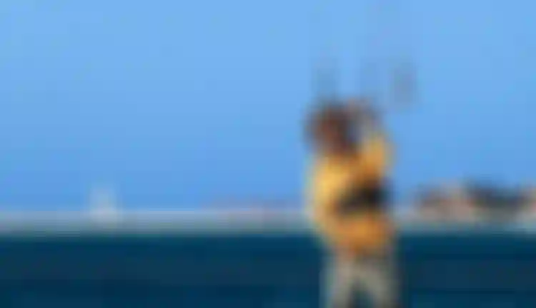 Smiling man in yellow shirt holding kitesurfing bar near ocean with sandy beach and blue sky in background