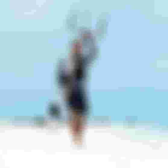 Kitesurfer walking barefoot on white sand beach, carrying a kiteboard and holding kite bar overhead under clear blue sky.