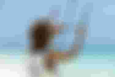 Woman with curly hair holding kitesurfing bar on a sunny beach, turquoise ocean in background, wearing a white surfing jersey.
