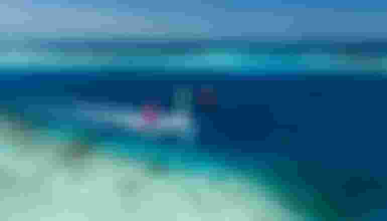 Aerial view of multiple colorful kite surfers gliding over crystal-clear turquoise tropical waters near a coral reef.
