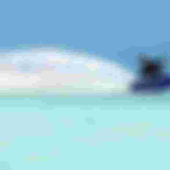 Man kitesurfing on a Duotone board in turquoise tropical waters, creating a large spray wake under a clear blue sky.
