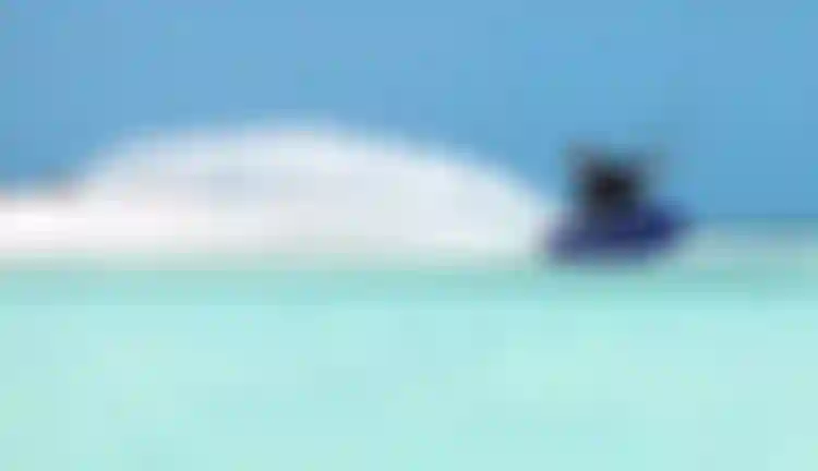 Man kitesurfing on a Duotone board in turquoise tropical waters, creating a large spray wake under a clear blue sky.