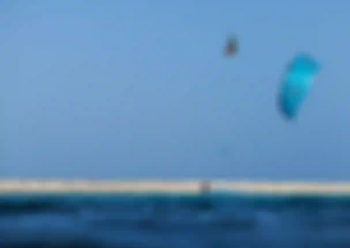 Kitesurfer riding turquoise ocean waters near a sandy shore, with a large blue kite and another rider airborne against a clear blue sky.
