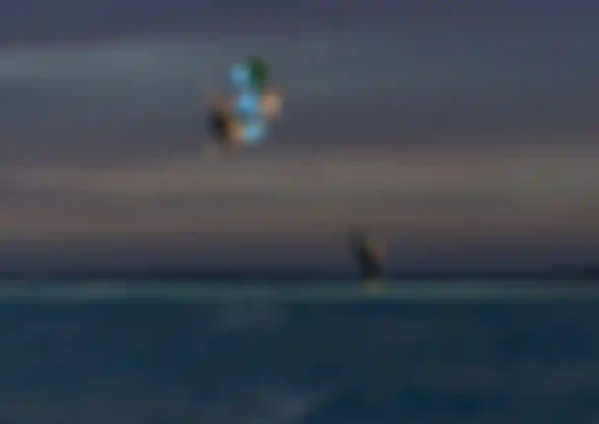 Two kitesurfers performing aerial tricks high above the ocean at dusk, while a person stands in shallow water watching below.