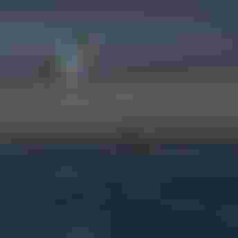 Two kitesurfers performing aerial tricks high above the ocean at dusk, while a person stands in shallow water watching below.