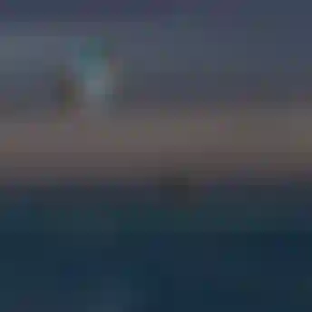 Two kitesurfers performing aerial tricks high above the ocean at dusk, while a person stands in shallow water watching below.