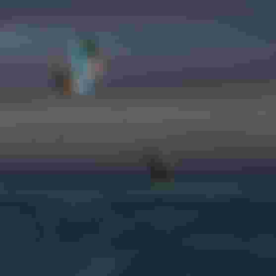 Two kitesurfers performing aerial tricks high above the ocean at dusk, while a person stands in shallow water watching below.