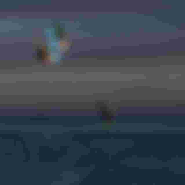 Two kitesurfers performing aerial tricks high above the ocean at dusk, while a person stands in shallow water watching below.