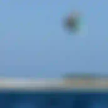 Kitesurfer performing a high aerial trick above turquoise ocean water near a sandy shoreline under a clear blue sky.