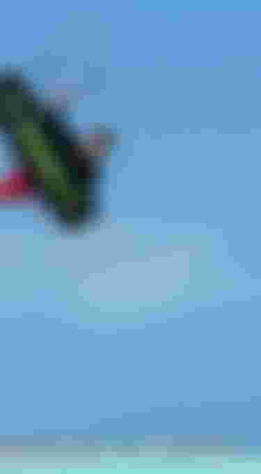 Kitesurfer mid-air grab holding a Duotone board above a turquoise tropical lagoon with white sand beach under clear blue sky.