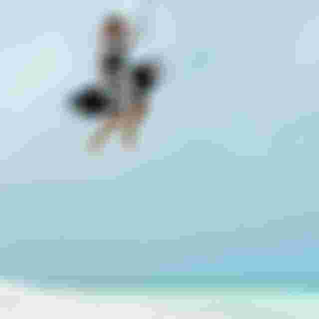 Male kitesurfer airborne mid-trick, holding kiteboard, over a white sand beach with turquoise water and cloudy sky.