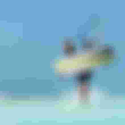 Male kitesurfer wading through shallow turquoise water holding a Duotone kiteboard and kite bar under a clear blue sky.
