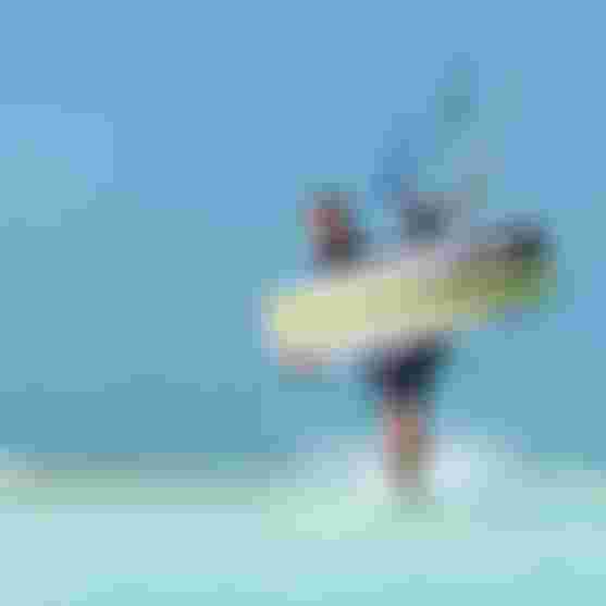 Male kitesurfer wading through shallow turquoise water holding a Duotone kiteboard and kite bar under a clear blue sky.