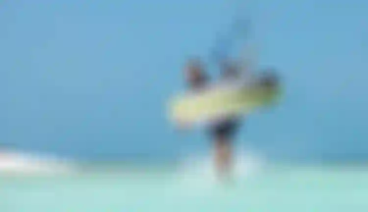 Male kitesurfer wading through shallow turquoise water holding a Duotone kiteboard and kite bar under a clear blue sky.