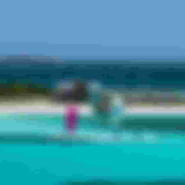 Three kitesurfers riding turquoise tropical waters near a white sand island with a beach house under clear blue skies.