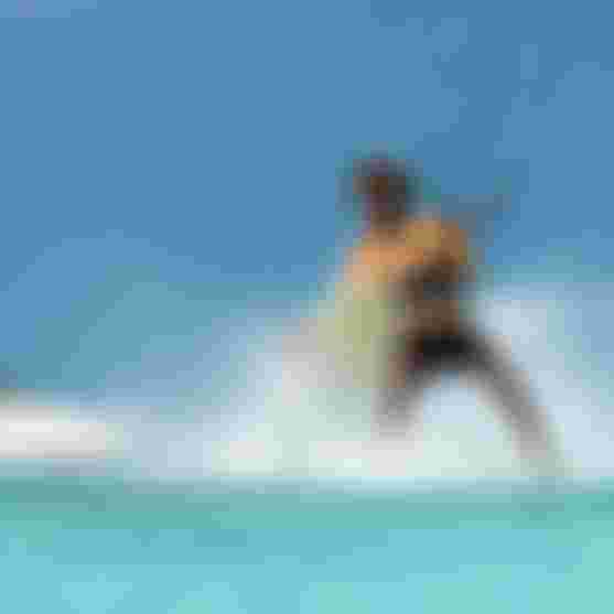 Male kitesurfer in orange shirt riding turquoise shallows near a white sand beach, holding a board mid-maneuver under clear blue sky.