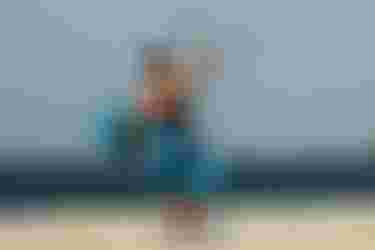 Male kitesurfer walking on a white sand beach, carrying a blue kiteboard and holding a kite control bar overhead.