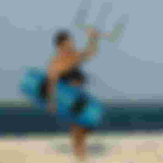 Male kitesurfer walking on a white sand beach, carrying a blue kiteboard and holding a kite control bar overhead.