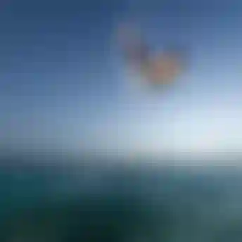 Kitesurfer in orange shirt launching aerial trick high above turquoise ocean water under clear blue sky with water droplets mid-air.