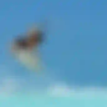 Kitesurfer in orange rashguard launching aerial trick above turquoise water against clear blue sky, water spray below board.