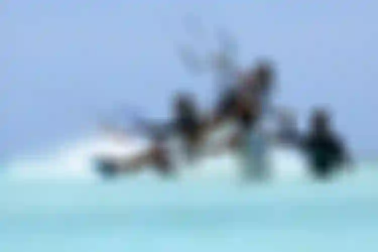 Group of kitesurfers riding through shallow turquoise water on a sunny day, with kite lines and colorful bars visible above.
