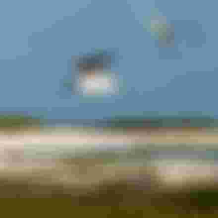 Kitesurfer airborne with a yellow-green kite over a coastal lagoon, seabirds in flight, white sandy shore and turquoise water below.
