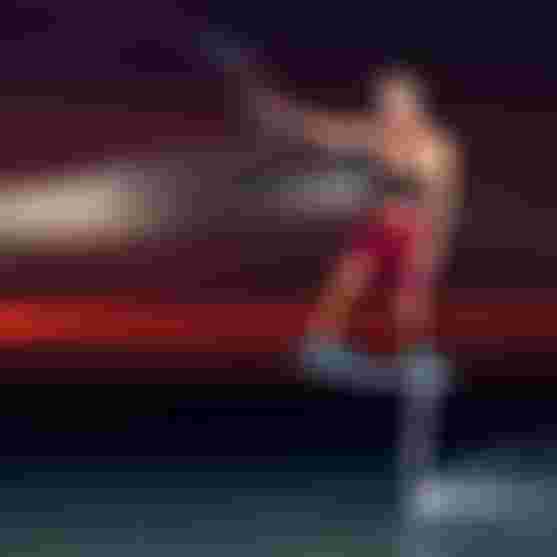 Man kite foiling above water at sunset, wearing red board shorts, holding kite bar while elevated on hydrofoil board against dramatic red sky.