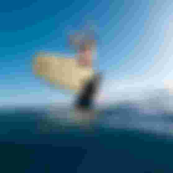 A man kiteboarding over the ocean, holding a kite bar, with a clear blue sky and water splashing around the board.