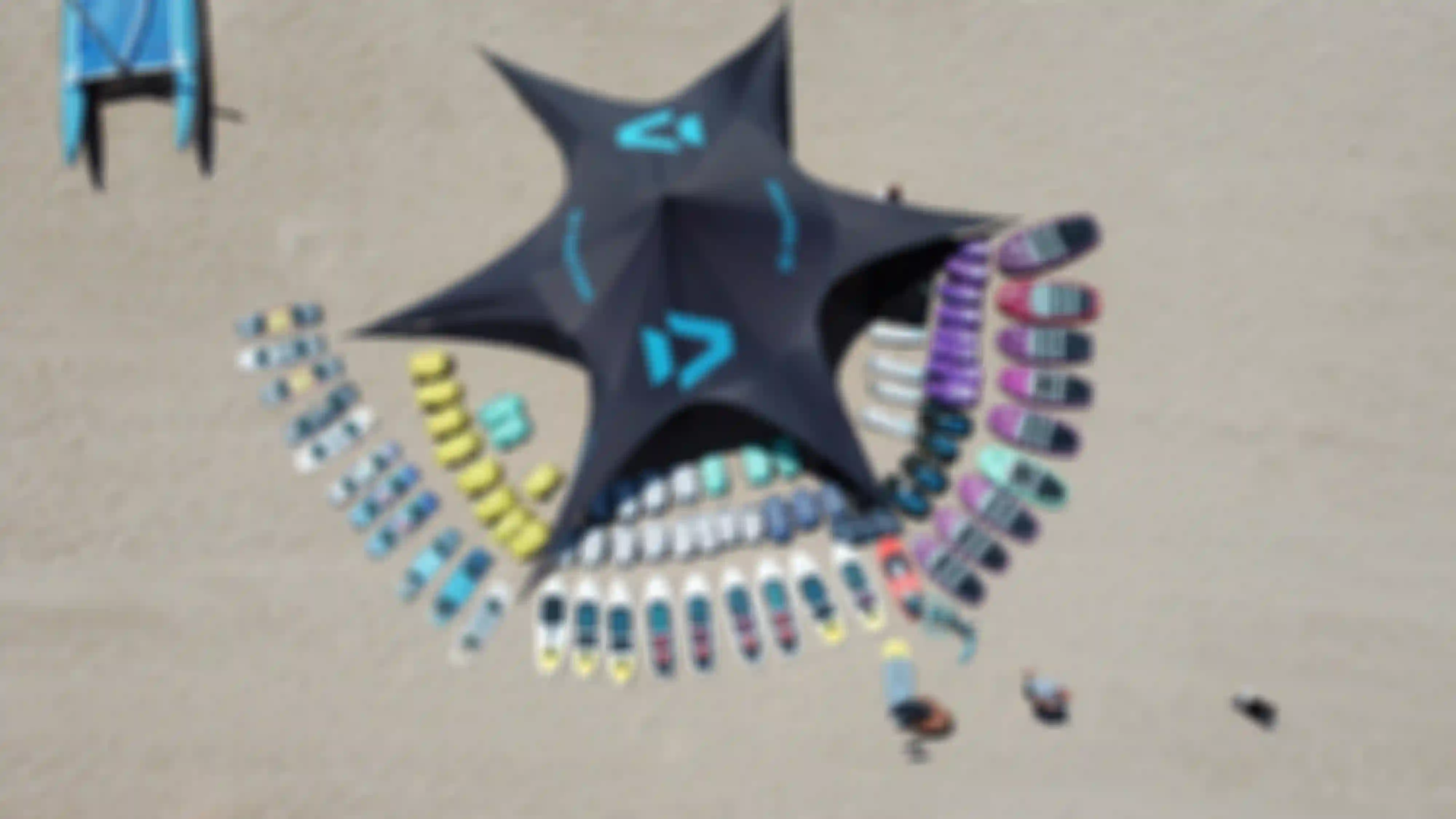Aerial view of a beach with a star-shaped tent labeled "Duotone," surrounded by colorful surfboards and gear arranged in semicircles on the sand.