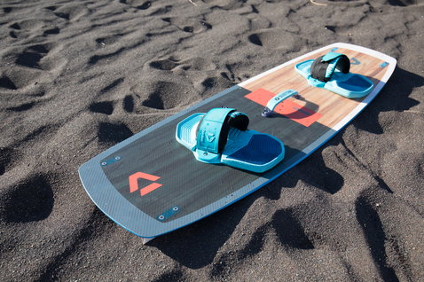 Duotone Gonzales twin-tip kiteboard in dark charcoal with teal, wood grain and coral accents on dark volcanic sand beach
