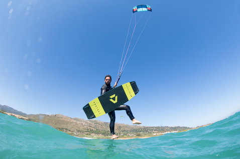 Duotone Select Textreme twin-tip kiteboard durante il kitesurf su una spiaggia oceanica con montagne sullo sfondo