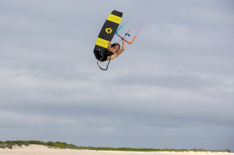 Duotone Select Textreme twin-tip kiteboard durante il kitesurf su una spiaggia costiera con dune e vegetazione