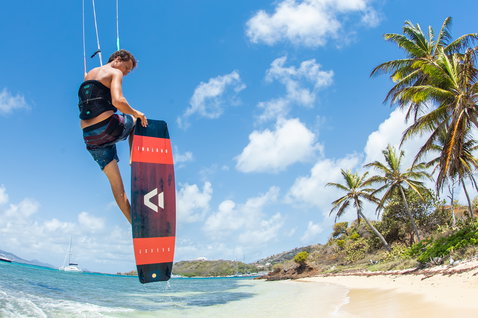 Duotone Select kiteboard twin-tip durante il kitesurf in spiaggia tropicale con palme e oceano turchese