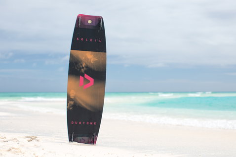 Duotone Soleil Twin-Tip Kiteboard in dunkel anthrazit schwarz mit orange und pink an einem tropischen Strand mit türkisem Wasser