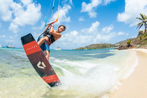 Duotone Trust Bar Quad Control Steuerbar beim Kitesurfen in einer tropischen Strandlagune mit Segelbooten und Palmen