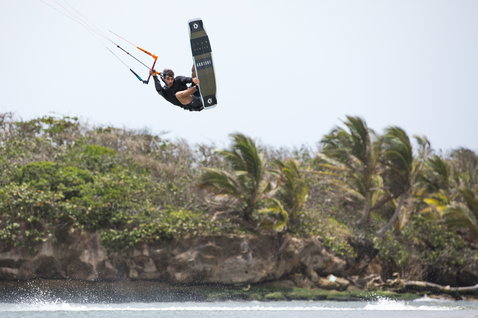 Duotone Team Series tabla de kitesurf twin-tip durante el kitesurf en una playa costera tropical con palmeras y costa rocosa