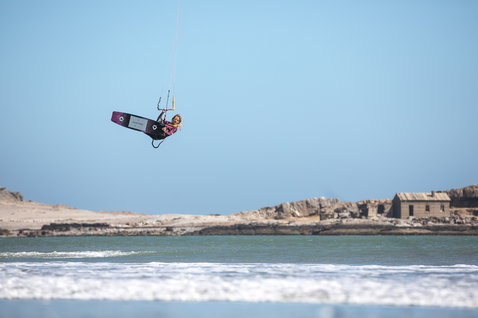 Duotone Soleil Textreme twin-tip kiteboard while kitesurfing at a coastal beach with rocky shoreline and turquoise water