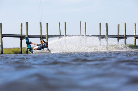 Duotone Gambler 2020 wake-style kiteboard durante il kitesurf in una laguna costiera con molo in legno ed erbe di palude