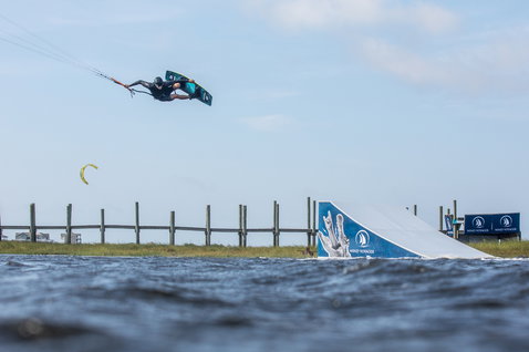 Duotone Gambler wake-style kiteboard durante il wakestyle su laguna di acque piatte con molo in legno e riva erbosa